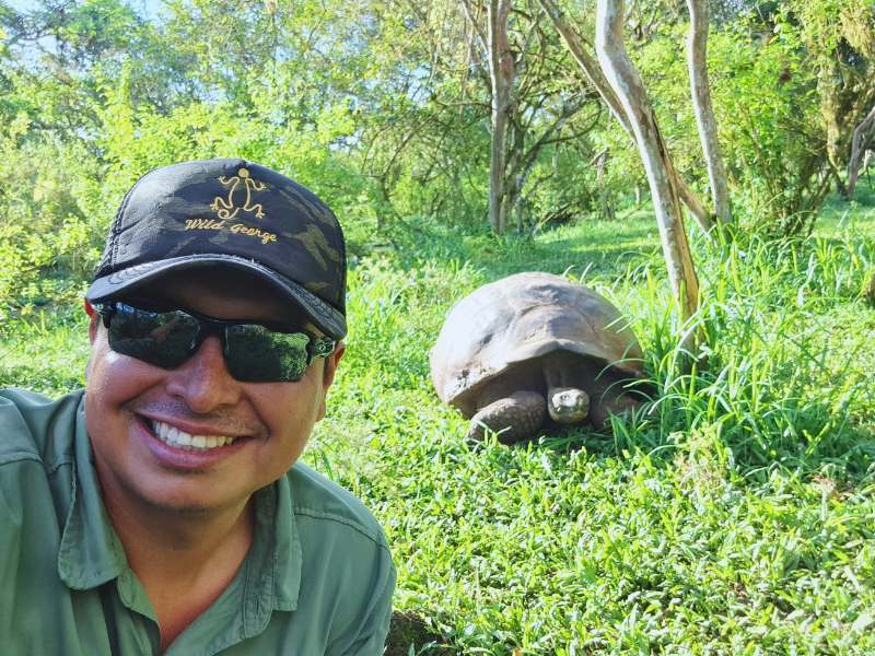 Wild George smiling with a Galápagos tortoise in a lush green environment.